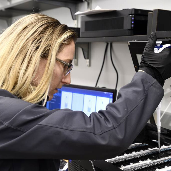 Photo of blonde, female researcher wearing a black flannel jacket and glasses holding a pipette.